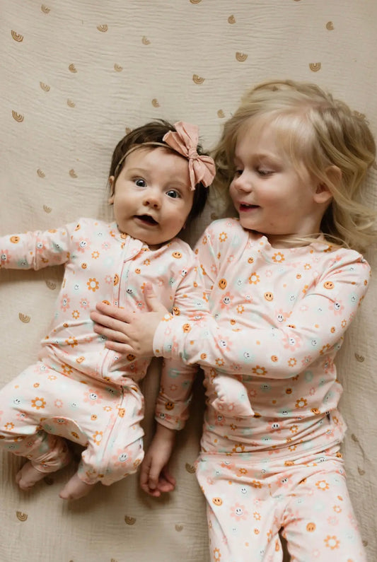 baby girl and toddler girl wearing matching baby bamboo pajama and toddler bamboo pajama set in pink with daisies and smiley faces